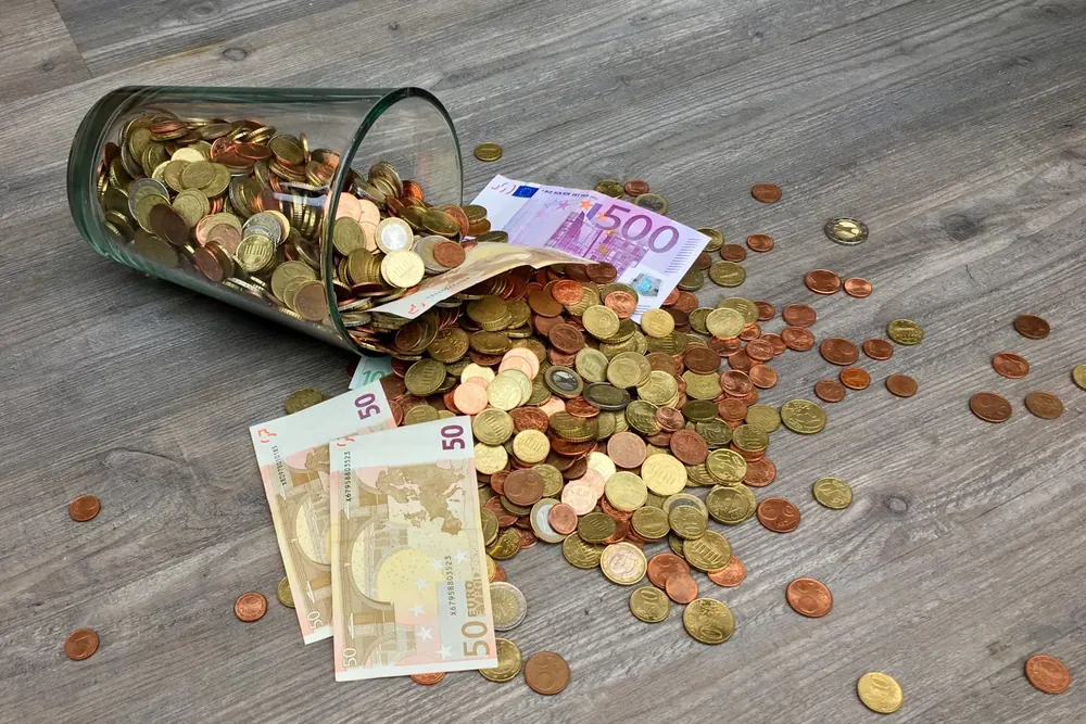 Bote de cristal volcado con monedas y billetes de euro esparcidos sobre una mesa de madera